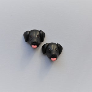 K&ouml;nnte beinhalten: Zwei schwarze Hundekopf-Figuren mit rosa Zungen. Die Hundek&ouml;pfe haben braune Augen und Schlappohren. Die Figuren befinden sich auf einem hellgrauen Hintergrund.
