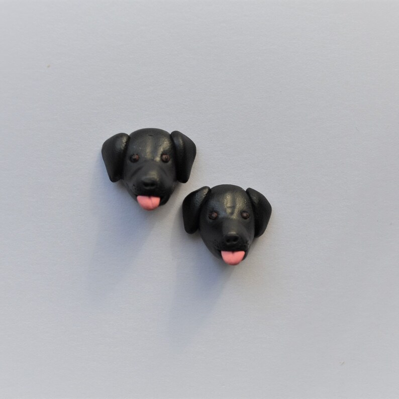 K&ouml;nnte beinhalten: Zwei schwarze Hundekopf-Figuren mit rosa Zungen. Die Hundek&ouml;pfe haben braune Augen und Schlappohren. Die Figuren befinden sich auf einem hellgrauen Hintergrund.