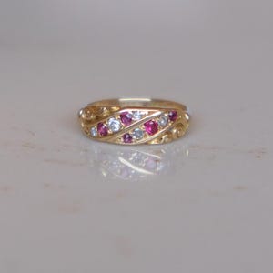 May include: A gold ring with a repeating pattern of alternating red and white gemstones. The ring is set on a white surface. The gemstones are small and round, and the ring has a textured band.