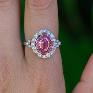 Puede incluir: Un anillo con una piedra preciosa rosa ovalada central rodeada por un halo de piedras rojas más pequeñas, encerrado en un halo exterior más grande de piedras blancas transparentes en una banda de metal plateado con diseño de brazo dividido.