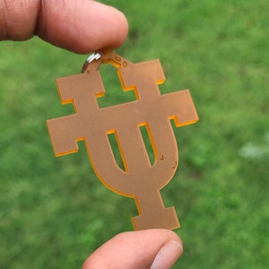 May include: A translucent orange keychain with a stylized letter or symbol design. The keychain is held up against a blurred green background. The symbol has a unique shape with a top bar and a bottom bar.