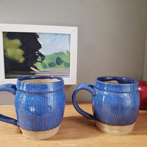 Peut inclure: Deux tasses en céramique bleues avec des bords blancs et un glaçage moucheté. Les tasses ont une forme arrondie et une poignée sur le côté. Les tasses sont posées sur une surface en bois.