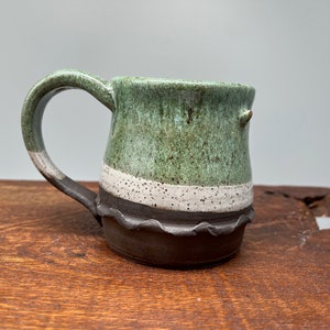 May include: A small, green ceramic mug with a white band and a brown rim. The mug has a speckled glaze and a slightly uneven shape.