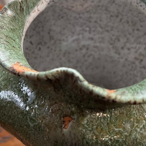 May include: A green ceramic vase with a textured, speckled glaze. The vase has a wide, rounded opening and a slightly irregular rim.