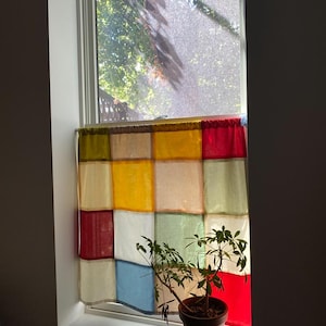 Pode incluir: Uma cortina colorida de retalhos está pendurada numa janela, com quadrados vermelhos, amarelos, azuis e bege. Uma planta em vaso está na janela em frente à cortina. A janela dá para uma árvore.