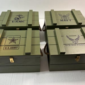 May include: Four olive green wooden storage boxes with hinged lids. Each box features a different U.S. military branch emblem and text: USMC, NAVY, U.S. ARMY, and U.S. AIR FORCE. The boxes have metal clasps and rope handles.