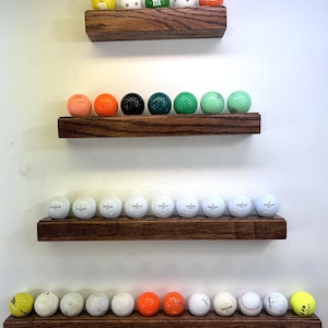 May include: Four wooden shelves with golf balls displayed on them. The top shelf has five golf balls with different designs, the second shelf has eight golf balls in various colors, the third shelf has 12 white golf balls, and the bottom shelf has 12 golf balls in various colors.