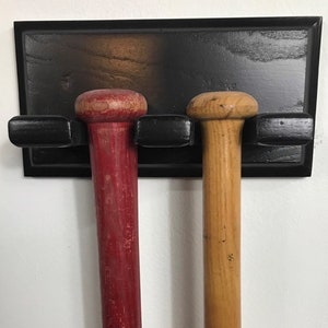 May include: A black wooden baseball bat rack with two bats, one red and one natural wood, mounted on a wall.