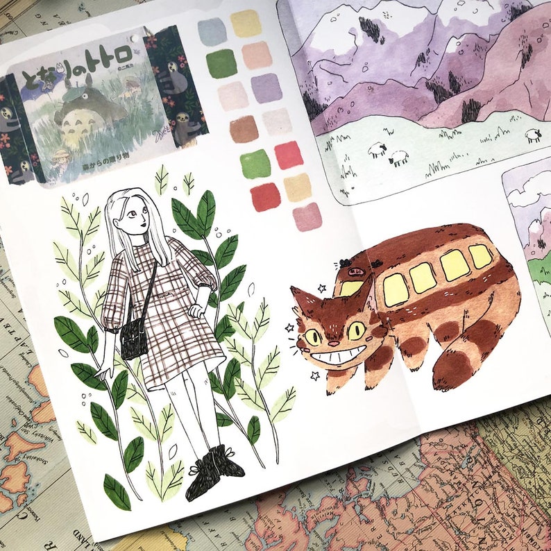 Puede incluir: Un cuaderno abierto con ilustraciones y muestras de color. Un dibujo presenta a una chica con un vestido a cuadros rodeada de hojas. Otro muestra un gato con forma de autob&uacute;s y una escena de monta&ntilde;a. Se ve texto japon&eacute;s.