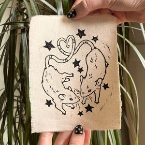 May include: A beige fabric patch featuring a black linocut design of two rats with intertwined tails forming a heart shape. Black star shapes are scattered around the rats. The patch is held up by two hands.