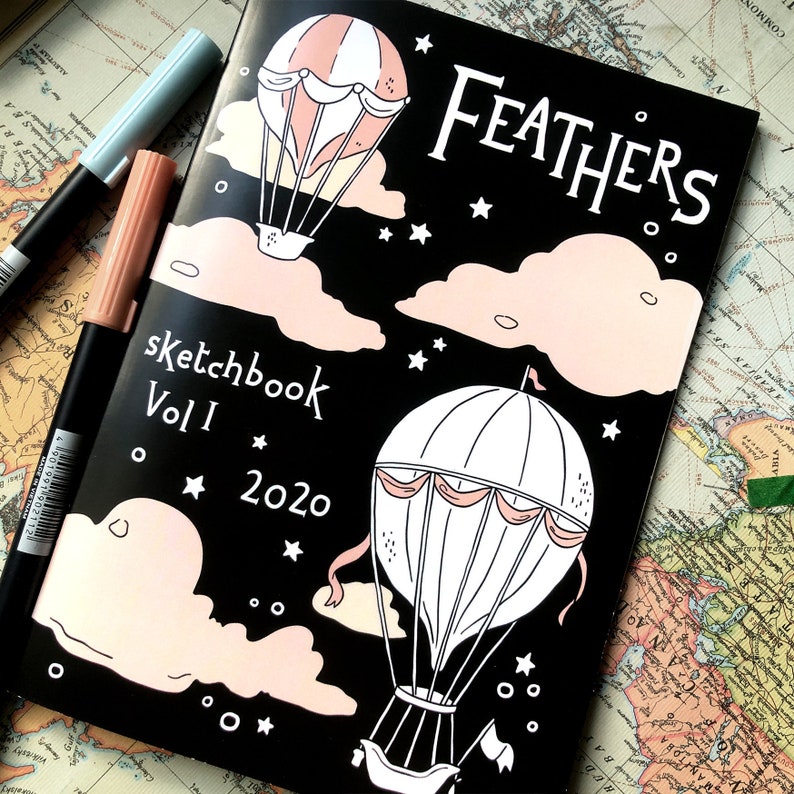 Puede incluir: Un cuaderno de bocetos negro titulado "Feathers" con un dise&ntilde;o de globo aerost&aacute;tico en blanco y rosa. La portada tambi&eacute;n presenta el texto "sketchbook Vol I 2020". Dos rotuladores en azul claro y melocot&oacute;n est&aacute;n a la izquierda.