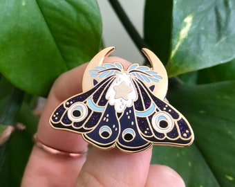 May include: A gold-tone enamel pin featuring a black and blue moth with a white star and crescent moon design. The moth has a glittery finish.