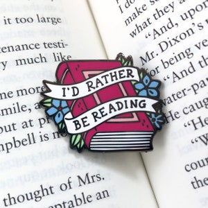 May include: A red enamel pin featuring a book design with the text "I'd Rather Be Reading". The pin has a blue and green floral design around the book.