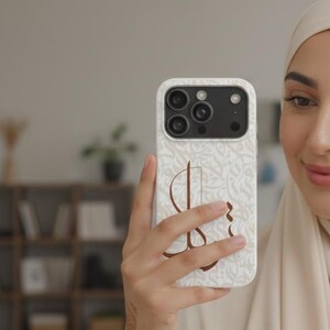 May include: A white smartphone case featuring a repeating light beige Arabic calligraphy pattern across the background and a large, prominent dark brown Arabic letter centered on the back. The case has a cutout for a triple-lens camera system.