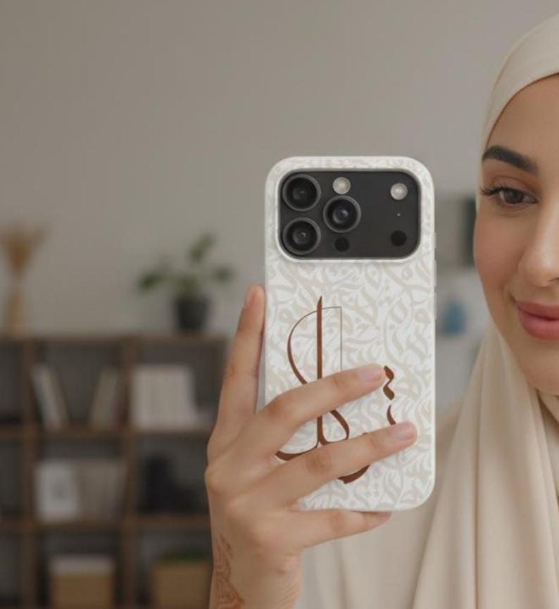 May include: A white smartphone case featuring a repeating light beige Arabic calligraphy pattern across the background and a large, prominent dark brown Arabic letter centered on the back. The case has a cutout for a triple-lens camera system.