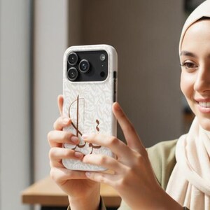 May include: A white smartphone case featuring a subtle, repeating light gray pattern and a prominent brown Arabic calligraphy design on the back. The case has a black camera module cutout with three circular lenses and a flash, fitting a modern smartphone.