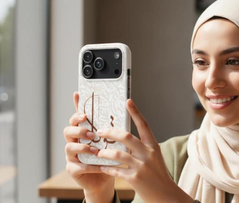 May include: A white smartphone case featuring a subtle, repeating light gray pattern and a prominent brown Arabic calligraphy design on the back. The case has a black camera module cutout with three circular lenses and a flash, fitting a modern smartphone.
