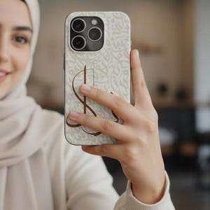 May include: A smartphone case featuring a light beige background with a subtle, repeating off-white calligraphic pattern. A prominent, dark brown stylized letter S is printed in the center of the case, which has a triple-lens camera cutout.