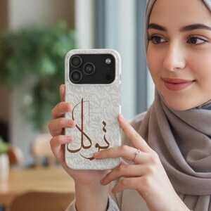 May include: A smartphone case featuring a light beige background with a subtle, repeating pattern of intricate, swirling script. Centered on the case is a large, bold, dark brown Arabic calligraphy design that stands out against the lighter backdrop.