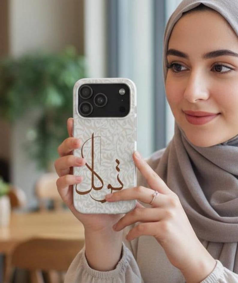 May include: A smartphone case featuring a light beige background with a subtle, repeating pattern of intricate, swirling script. Centered on the case is a large, bold, dark brown Arabic calligraphy design that stands out against the lighter backdrop.