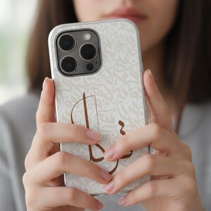 May include: A smartphone case featuring a light cream background with a subtle, repeating white floral pattern and a prominent, stylized brown Arabic calligraphic design centered on the back. The case has a cutout for a triple-lens camera system.