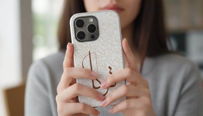 May include: A smartphone case featuring a light cream background with a subtle, repeating white floral pattern and a prominent, stylized brown Arabic calligraphic design centered on the back. The case has a cutout for a triple-lens camera system.
