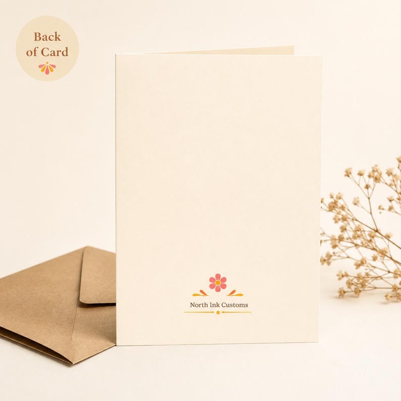 May include: A cream-colored greeting card standing upright with a brown kraft paper envelope to its left. The bottom center of the card features a small pink flower icon with orange accents and the text North Ink Customs in brown lettering.