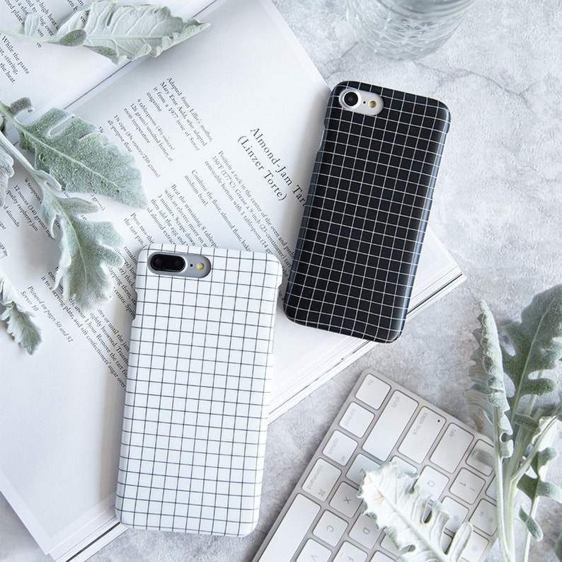 May include: Two black and white grid patterned phone cases. The cases are made for an iPhone and have a black and white grid pattern. The cases are on a white surface with a keyboard and a plant.