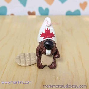 Peut inclure: Une petite figurine de castor brun portant un toque blanc avec une feuille d'érable rouge. Le castor a une petite queue brune et se tient sur une surface en bois.