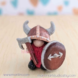 Peut inclure: Une petite figurine de gnome artisanale portant un casque brun et argent avec des cornes, une tunique rouge et tenant une hache argentée et un bouclier brun avec un motif de flèche argentée.