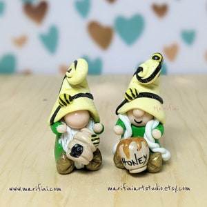 May include: Two miniature gnomes with yellow and black bee hats holding honey pots. The gnome on the left has a beard and the gnome on the right has a white scarf. Both gnomes are wearing green outfits.