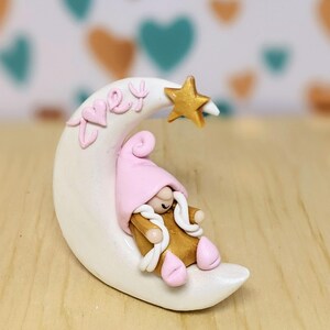 Könnte beinhalten: Eine skurrile Figur eines schlafenden Zwergs auf einem Halbmond. Der Zwerg trägt einen rosa Hut und goldfarbene Kleidung. Der Mond ist weiß mit dem Namen "Zoey" in Rosa. Ein goldener Stern sitzt über dem Mond.