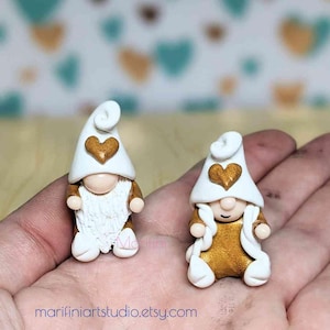 Può includere: Due figurine di gnomi bianche con dettagli dorati. Gli gnomi indossano cappelli bianchi con un cuore dorato sul davanti. Gli gnomi sono in piedi su una mano.