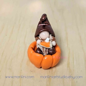 May include: A small gnome figurine made of polymer clay, wearing a brown hat and sitting inside a bright orange pumpkin. The gnome is holding a slice of pumpkin pie with a dollop of whipped cream.