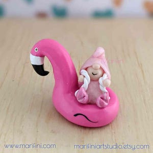 May include: A pink gnome figurine sitting on a pink inflatable flamingo. The gnome is wearing a pink hat and holding a white rope.