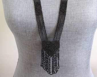 May include: A long black beaded necklace with a fringe pendant. The necklace is made of small black beads and has a V-shaped design.