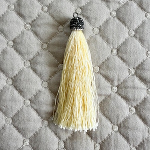 May include: A decorative tassel with a black beaded cap and a long, pale yellow fringe. The tassel is suspended against a neutral, quilted fabric background. The tassel is a craft supply.