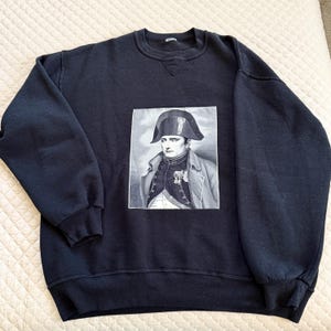 May include: A navy blue sweatshirt featuring a black and white portrait of Napoleon Bonaparte. The portrait is rectangular and centered on the front of the sweatshirt. The sweatshirt has a crew neck and long sleeves.