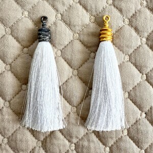 May include: Two white tassels with decorative metal caps. One cap is dark gray, the other is gold. The tassels are made of many strands of white thread, and are displayed on a beige quilted surface.