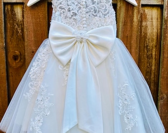 Puede incluir: Vestido formal blanco con un gran lazo y detalles de encaje. El vestido está en una percha blanca sobre un fondo de madera. El corpiño presenta encaje intrincado y pequeños botones, y la falda está en capas con tul.
