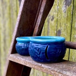 May include: Two blue and teal ceramic candle holders with a textured surface. The candle holders are on a wooden shelf.