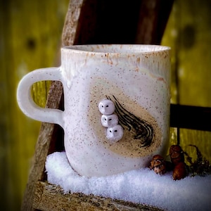 May include: White ceramic mug with a speckled glaze and a hand-painted design of three owls. The owls are white with black eyes and are arranged in a line. The mug is sitting on a wooden shelf with snow on it.