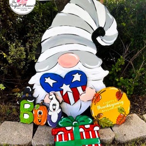 May include: A large wooden gnome with a white beard and a gray and white hat. The gnome holds a heart-shaped sign with stars and stripes. Additional wooden cutouts include a pumpkin, ghost, and a gift box. A sign says "Happy Thanksgiving".