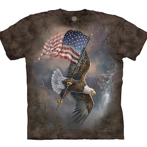 May include: A brown t-shirt with a tie-dye pattern features a bald eagle flying with its talons gripping an American flag. The eagle is flying over a mountain range with a waterfall in the background.