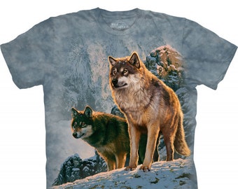 May include: A short-sleeved crew neck t-shirt in a mottled blue-grey color featuring a graphic print of two wolves standing on a snow-covered mountain slope. The wolves are depicted with detailed fur textures in shades of brown, grey, and white.