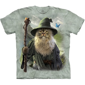 May include: A green t-shirt with a tie-dye effect featuring a cat dressed as a wizard with a long white beard, a pointed hat, and a staff. The cat is standing in a fantasy landscape with a blue butterfly flying above.