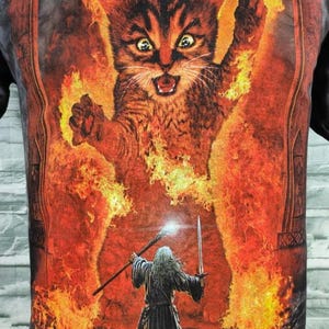 May include: Black t-shirt featuring a fiery cat graphic with a wizard holding a staff and sword. The cat is surrounded by flames, with a detailed illustration style. The design has a fantasy theme.