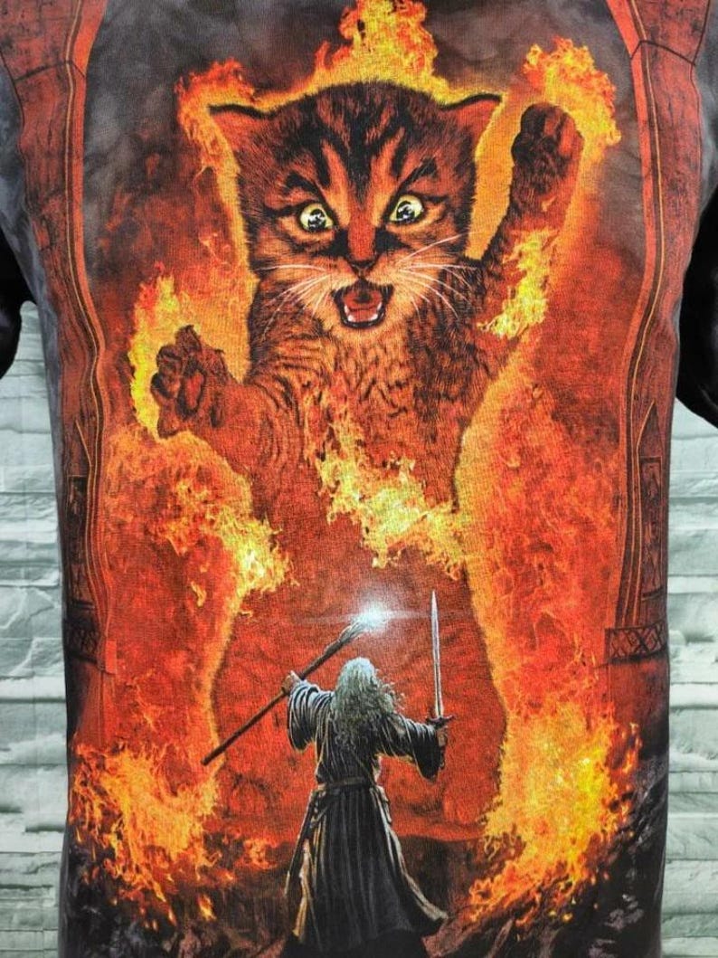 May include: Black t-shirt featuring a fiery cat graphic with a wizard holding a staff and sword. The cat is surrounded by flames, with a detailed illustration style. The design has a fantasy theme.