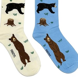 Puede incluir: Un par de calcetines con un diseño de oso y tocón de árbol. Un calcetín es blanco con un oso marrón y el otro es azul con un oso negro.