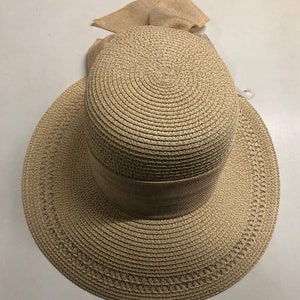 May include: A beige straw sun hat with a wide brim and a matching ribbon around the crown. The hat has a bow made of the same straw material on the top.